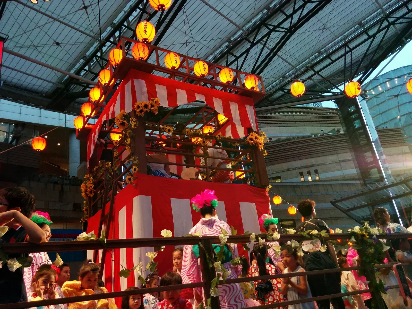東京麻布十番祭典會場，紅白帳棚下掛滿黃色燈籠，穿浴衣的大人與小孩站在欄杆旁。