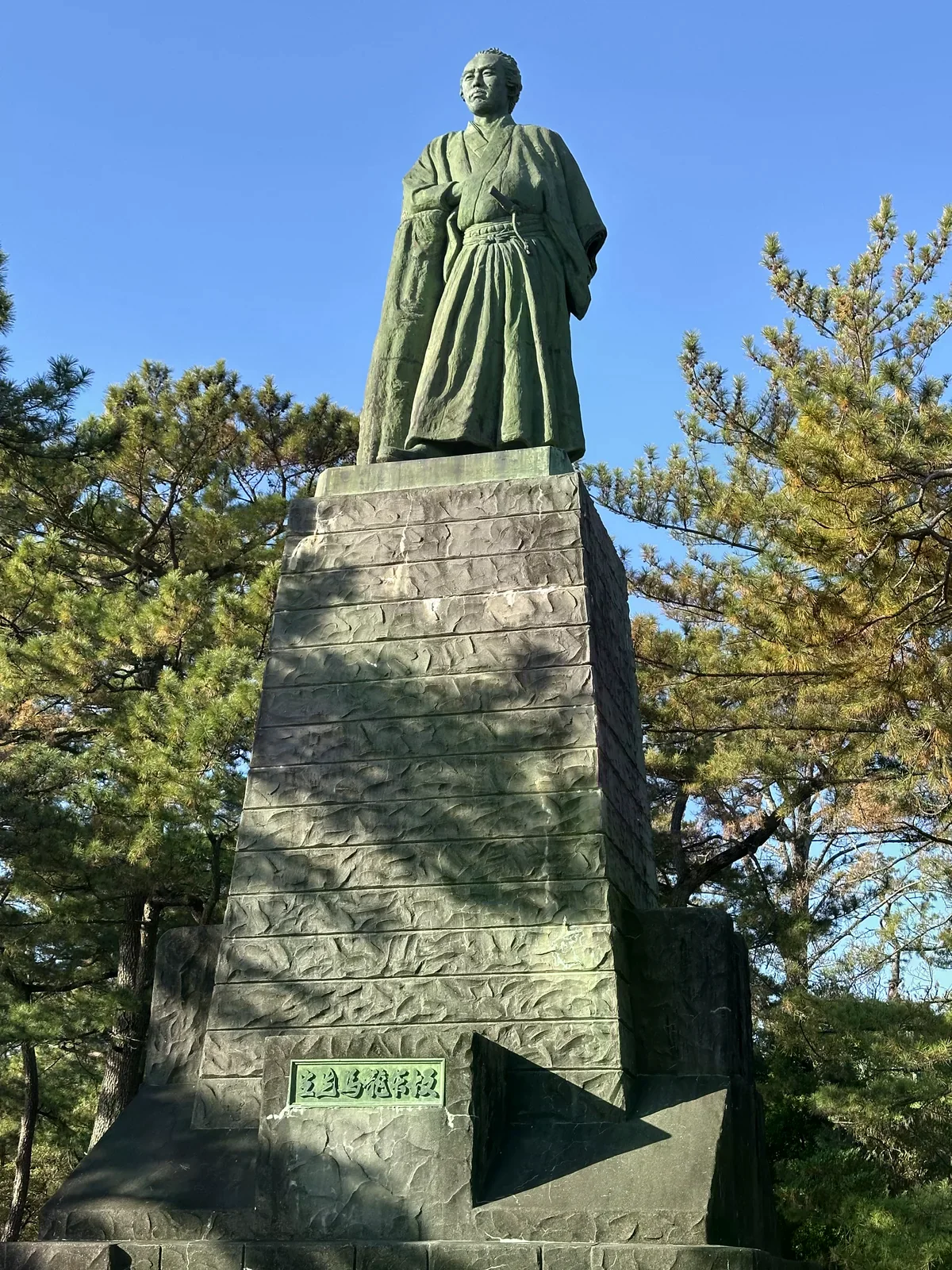 桂濱高石基座上的坂本龍馬青銅立像，背景是松樹與藍天