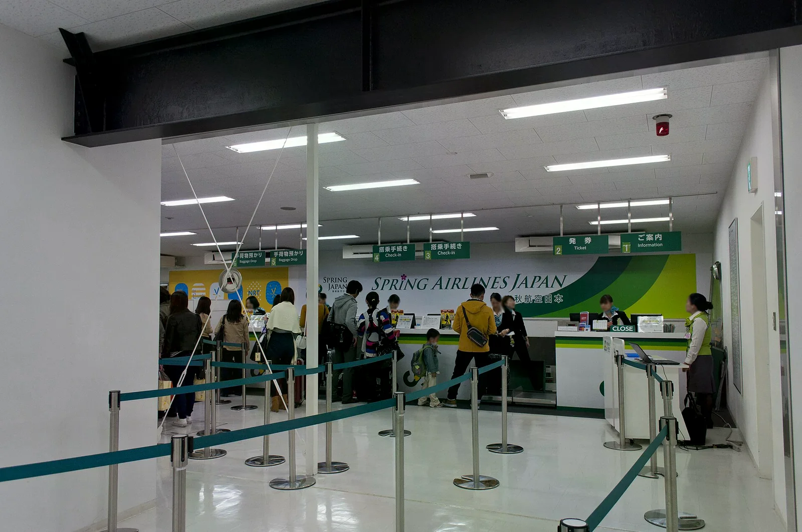 航空公司報到櫃台，綠色招牌寫著 SPRING AIRLINES JAPAN，旅客在藍色圍欄後排隊辦理報到