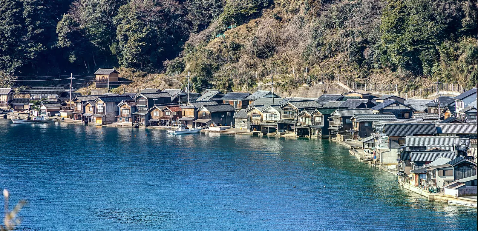 伊根灣岸一排黑色木造舟屋緊鄰平靜藍色海面，港邊停著小型漁船