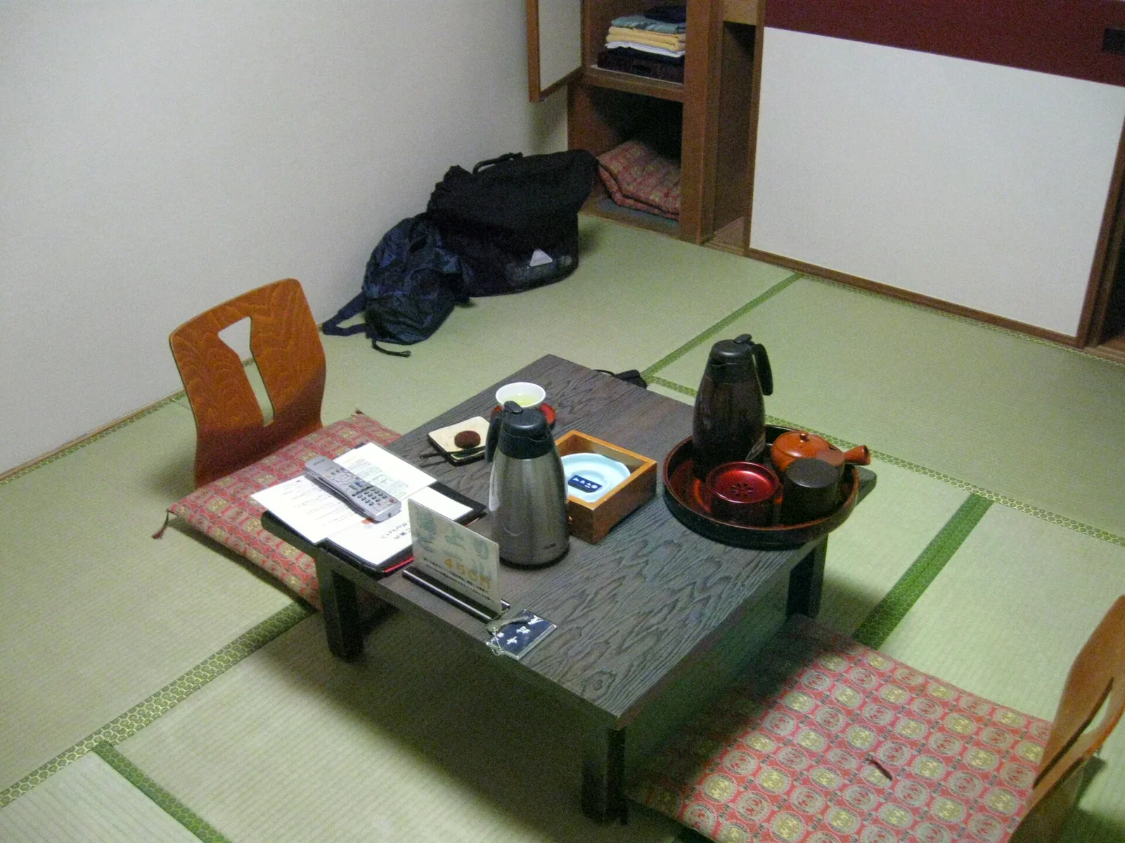 鋪著淺綠榻榻米的和室，中間一張矮木桌，桌上有保溫瓶、茶具托盤與鍵盤，旁邊放布椅墊與靠背椅，角落置物櫃堆了背包