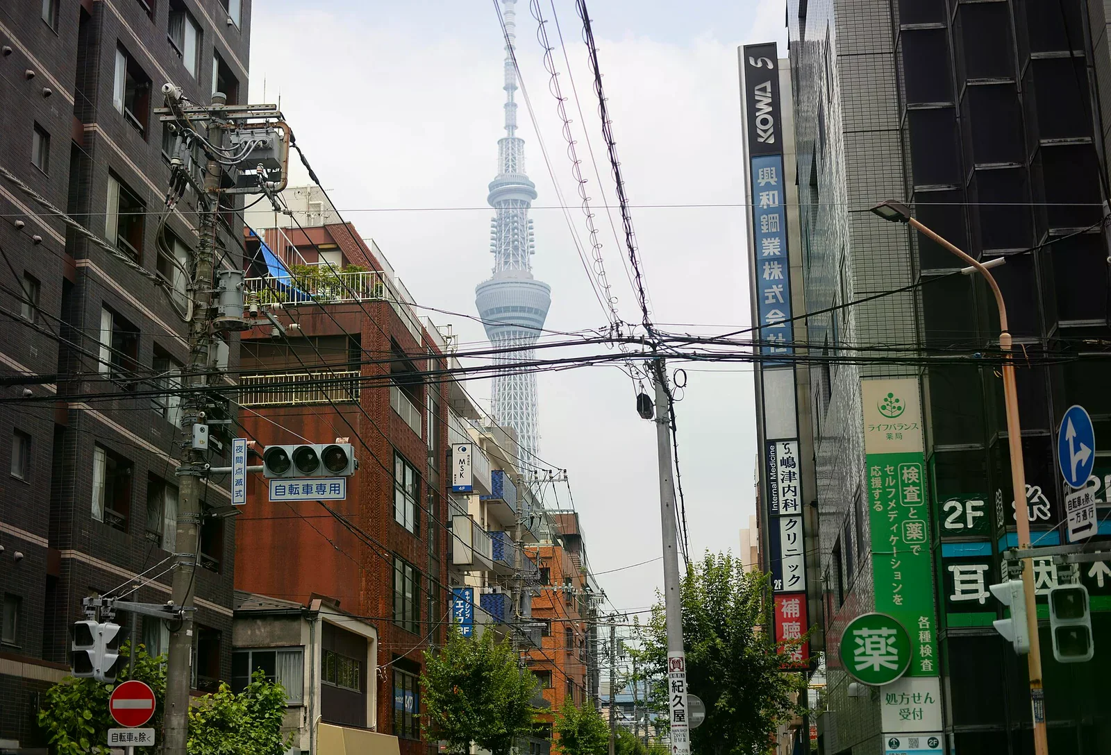東京晴空塔從住宅街高樓之間探出，前景是架空電線、路燈與兩側店家招牌
