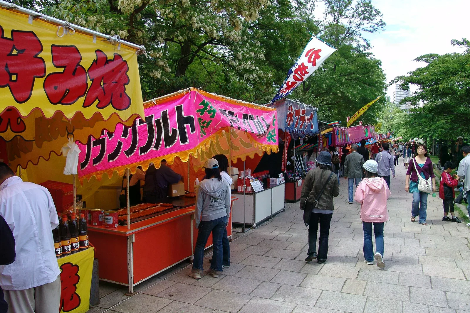 夏日祭典的屋台街，一整排黃色、粉紅色布幕攤位沿步道排列，人們在攤位前排隊購買小吃