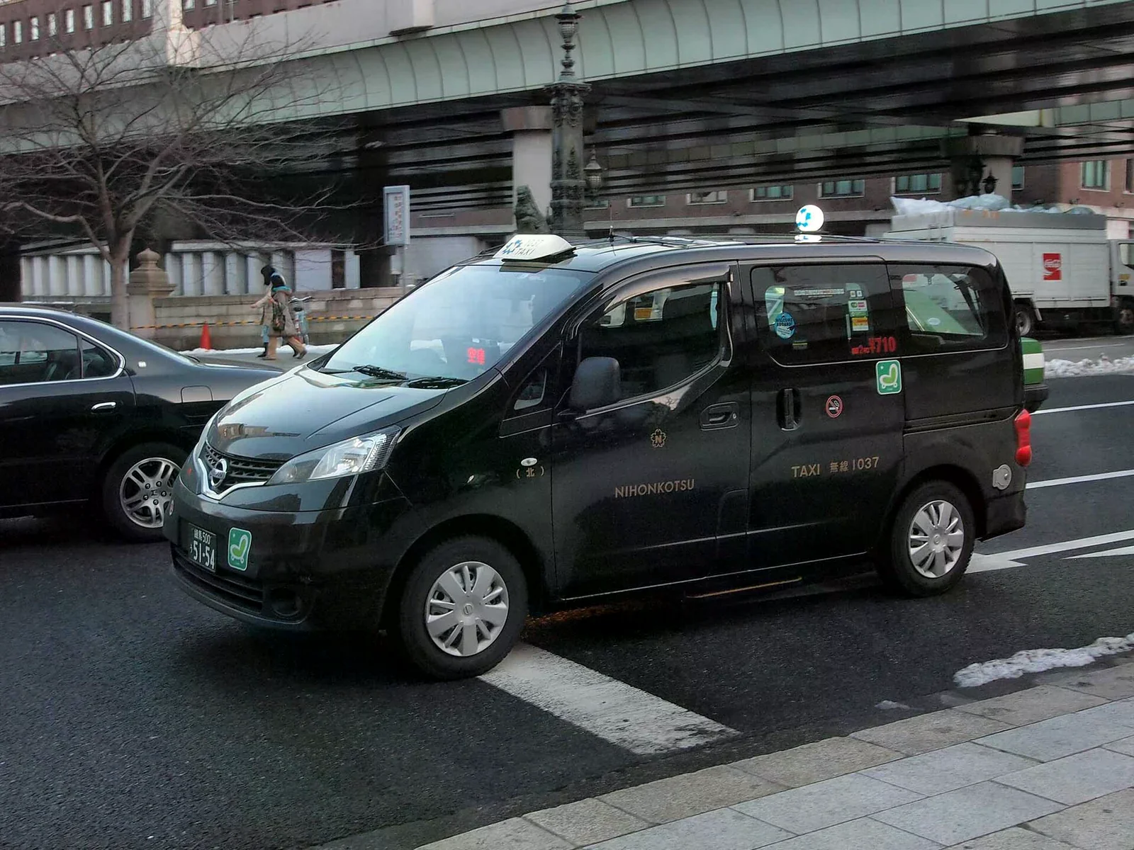 東京日本橋街邊一輛黑色日本交通 NV200 計程車，儀表板亮著空車標誌，車身停在路面。