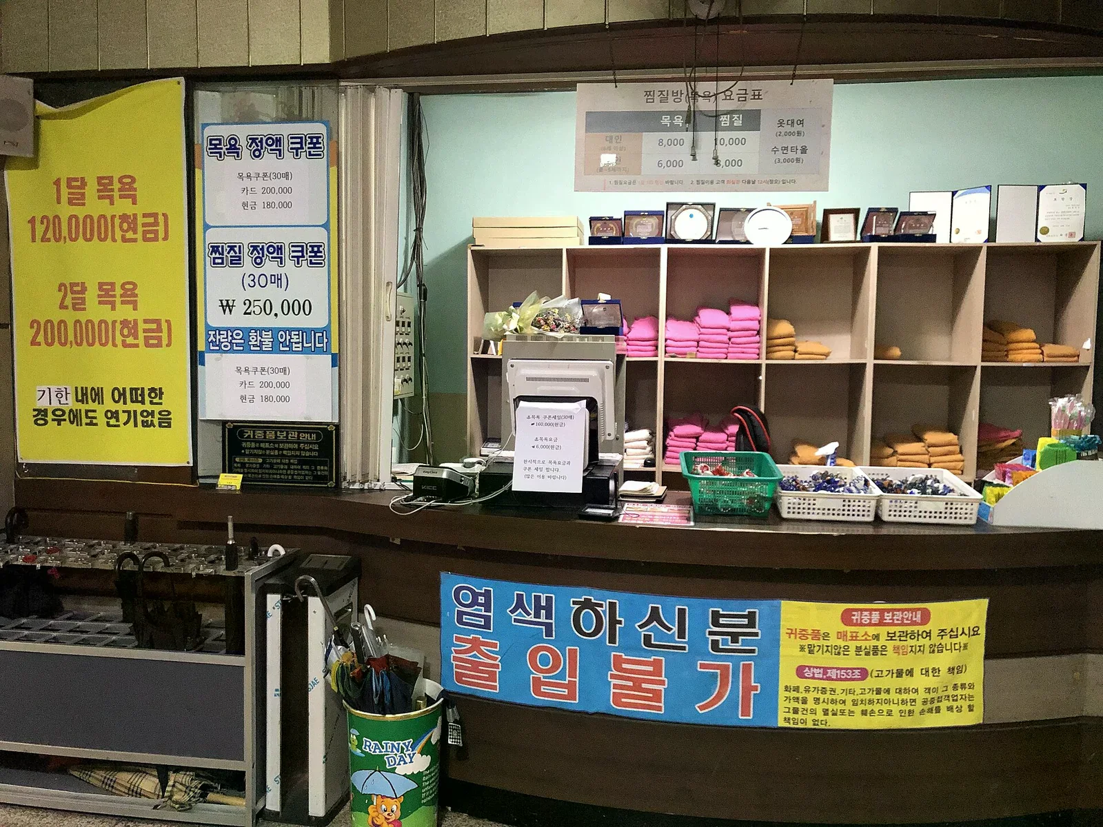 汗蒸幕櫃台，層架上疊放粉紅與米色的館內毛巾，桌上有收銀機、傘架與價目告示。