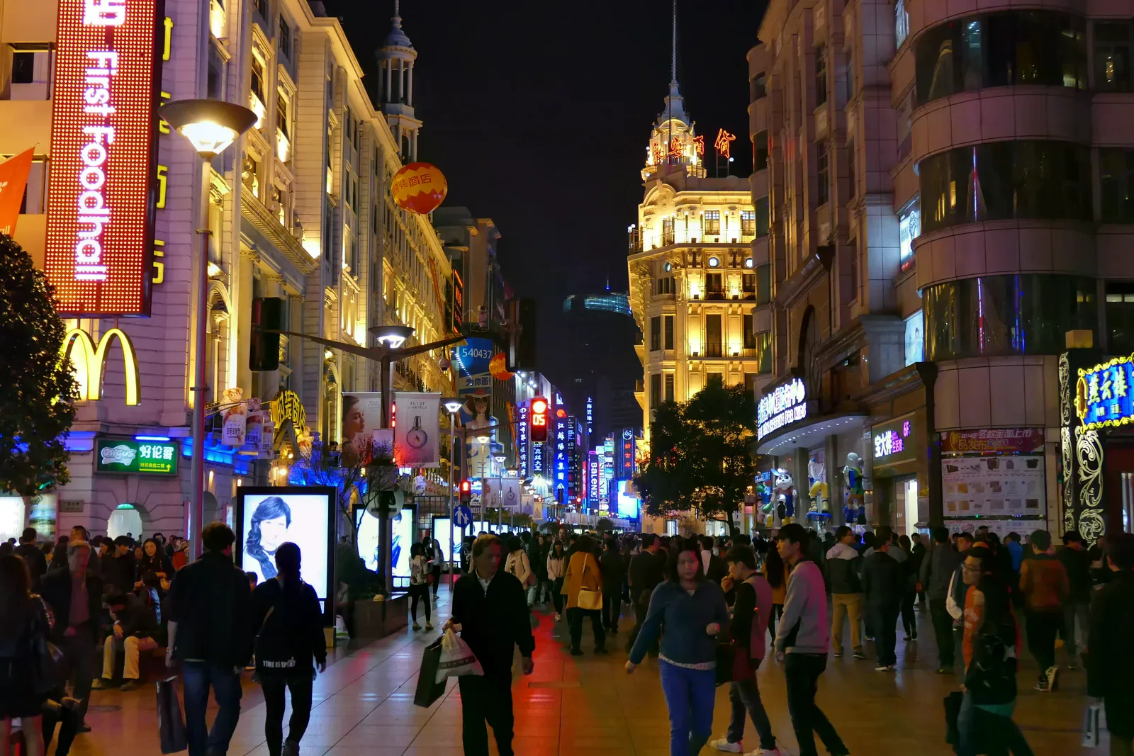 夜晚的南京路步行街上人潮往來，兩側百貨公司招牌亮燈，遠處可見裝飾藝術風格的永安百貨塔樓。