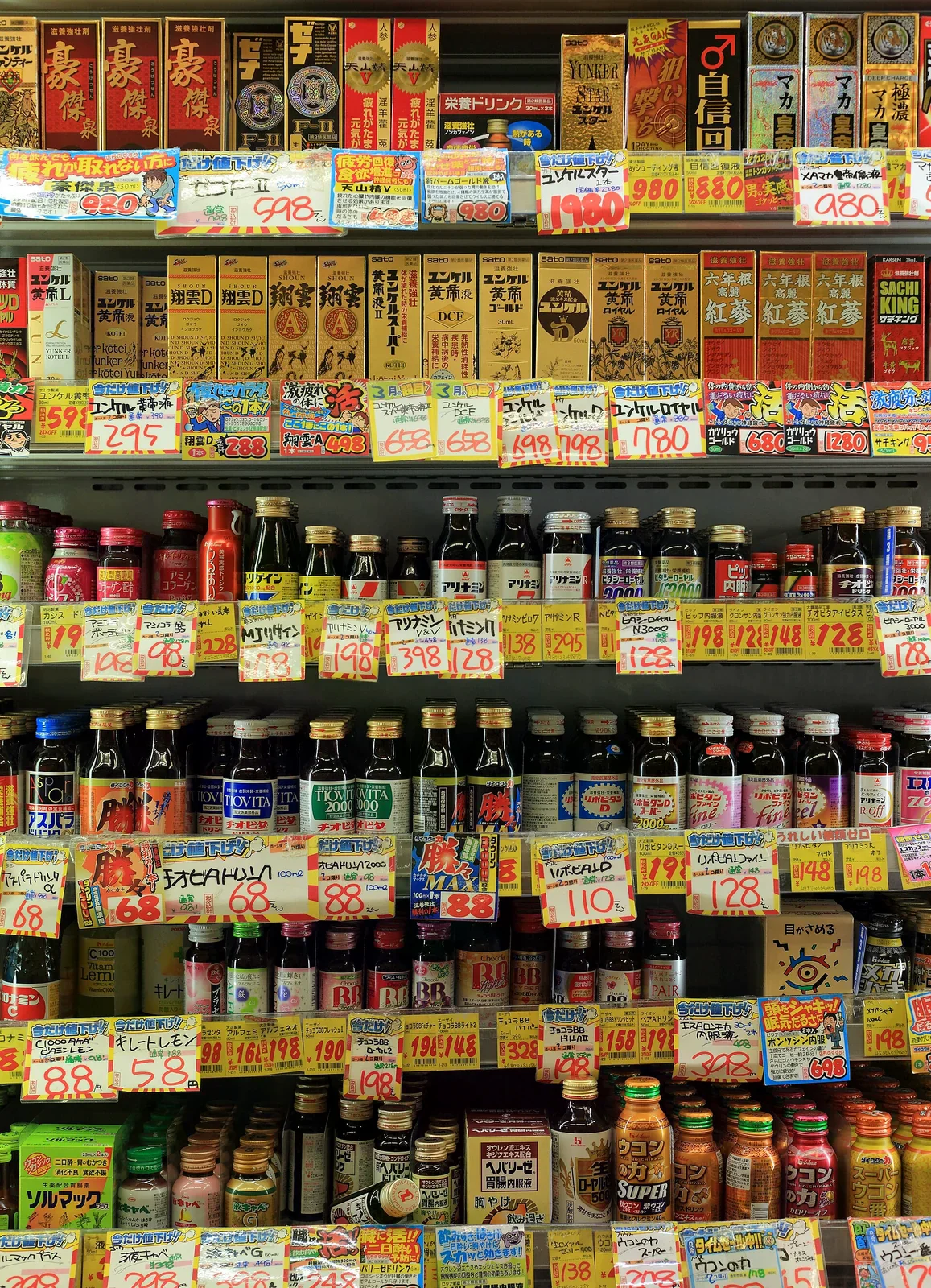 便利商店貨架上滿滿的營養飲料與補養品瓶罐，每層前方貼著彩色手寫風格價格標籤