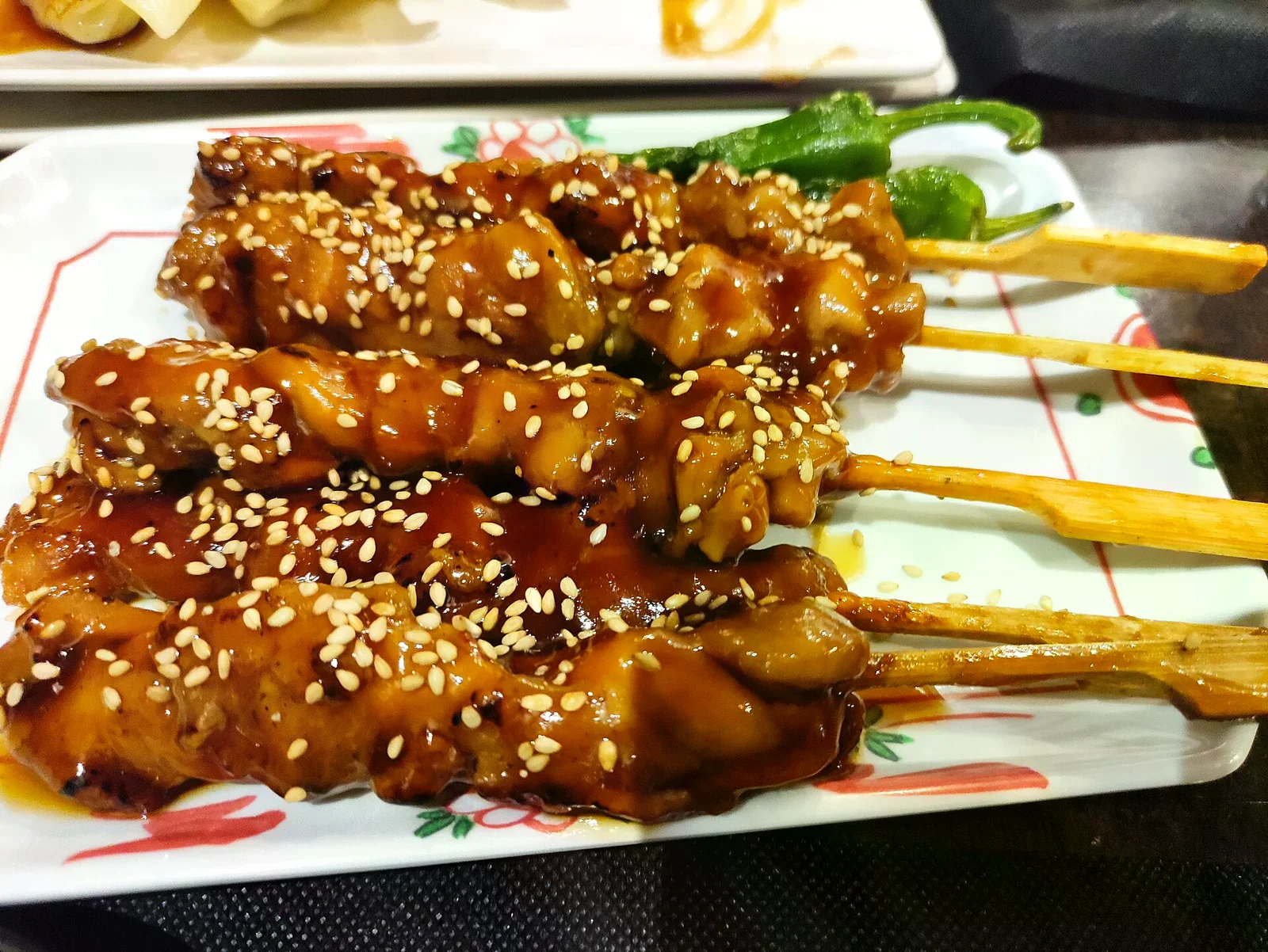 白盤上擺著四串照燒醬汁串烤雞肉，表面撒滿白芝麻，旁邊有兩根青椒