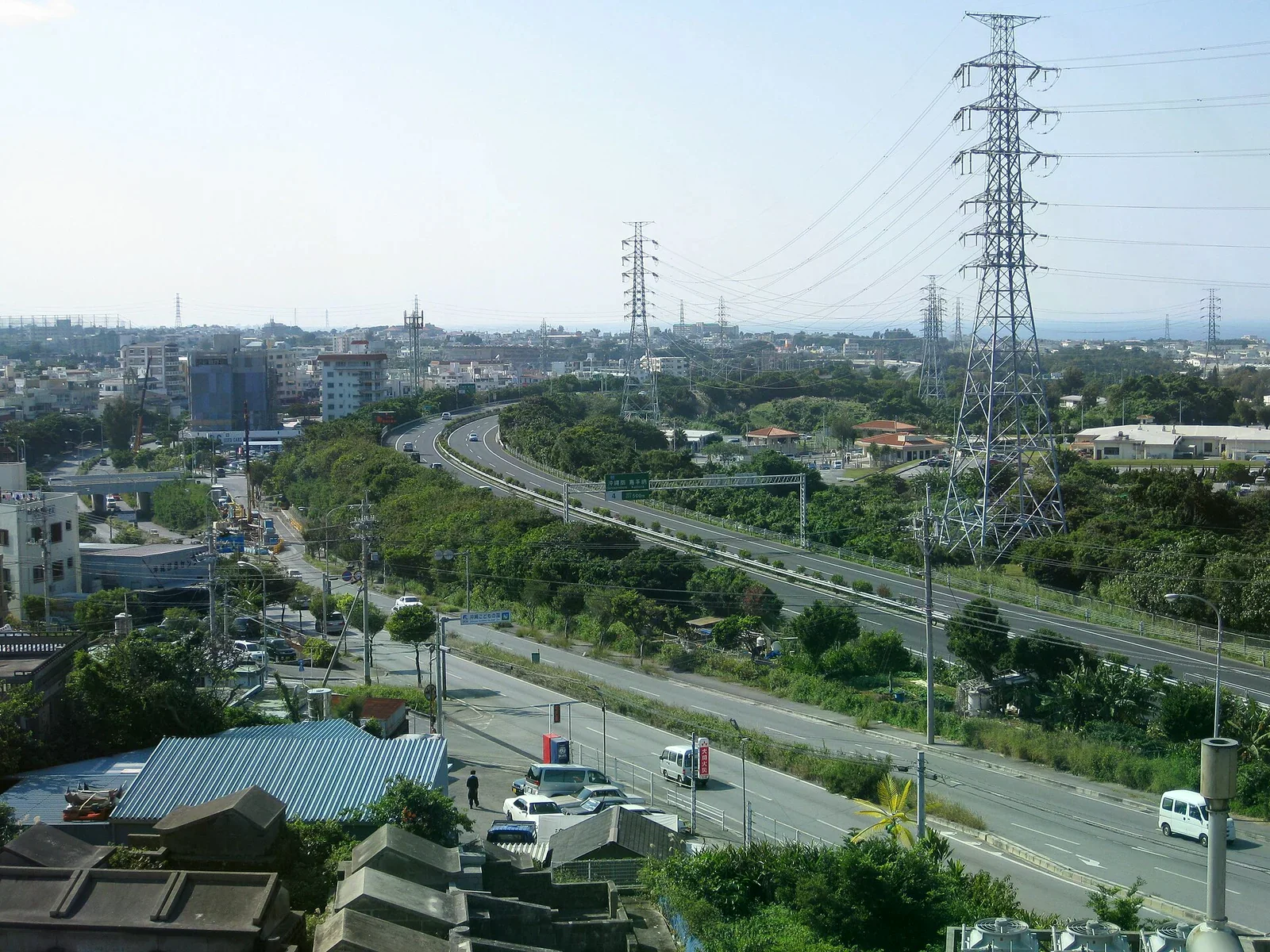 俯瞰沖繩市區一條多線道快速道路，路線右彎穿過綠地與低樓層建築，旁邊有高壓電塔