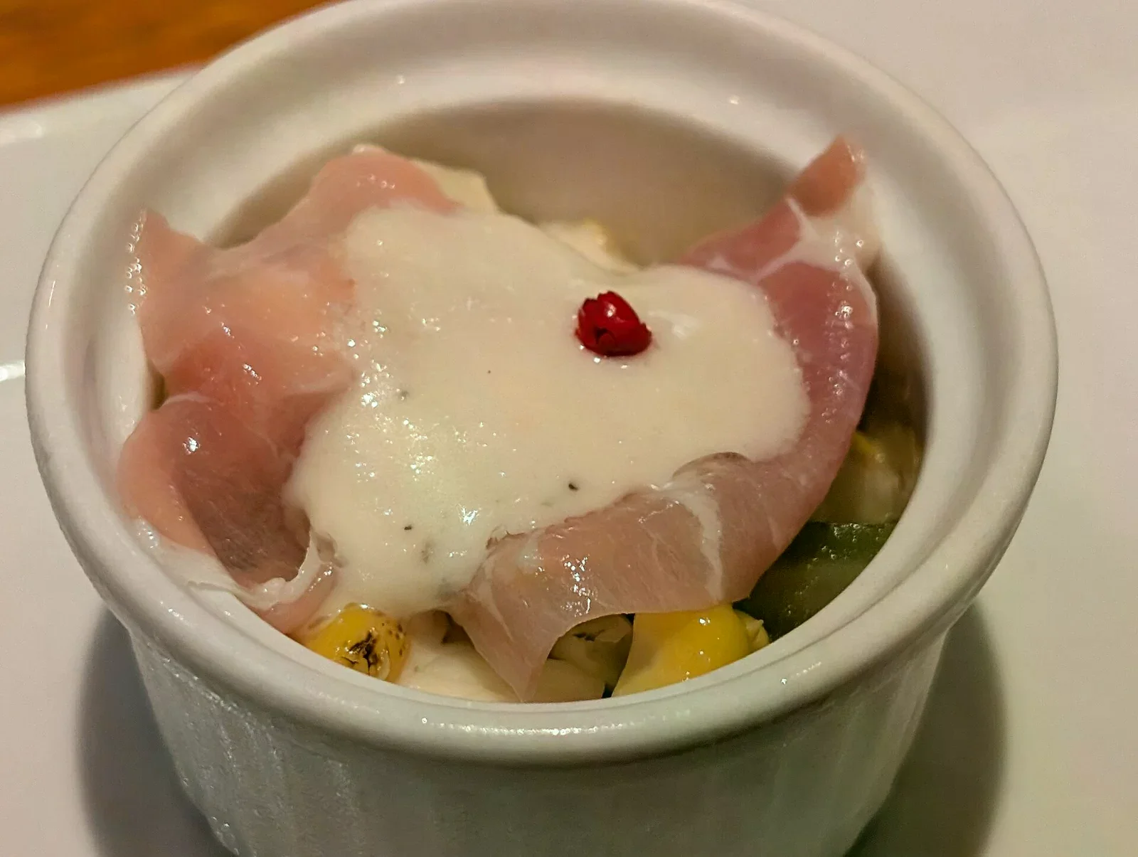 白色陶瓷烤盅裡裝著起司白醬料理，上面鋪了生火腿片，中央點綴一顆紅胡椒