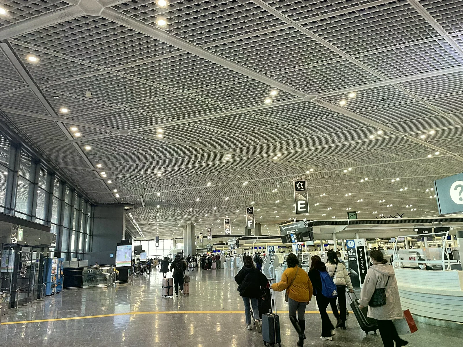 成田機場出境大廳內部，挑高格狀天花板下旅客拉著行李車走向報到櫃台，上方懸掛 E、F、G 登機門編號