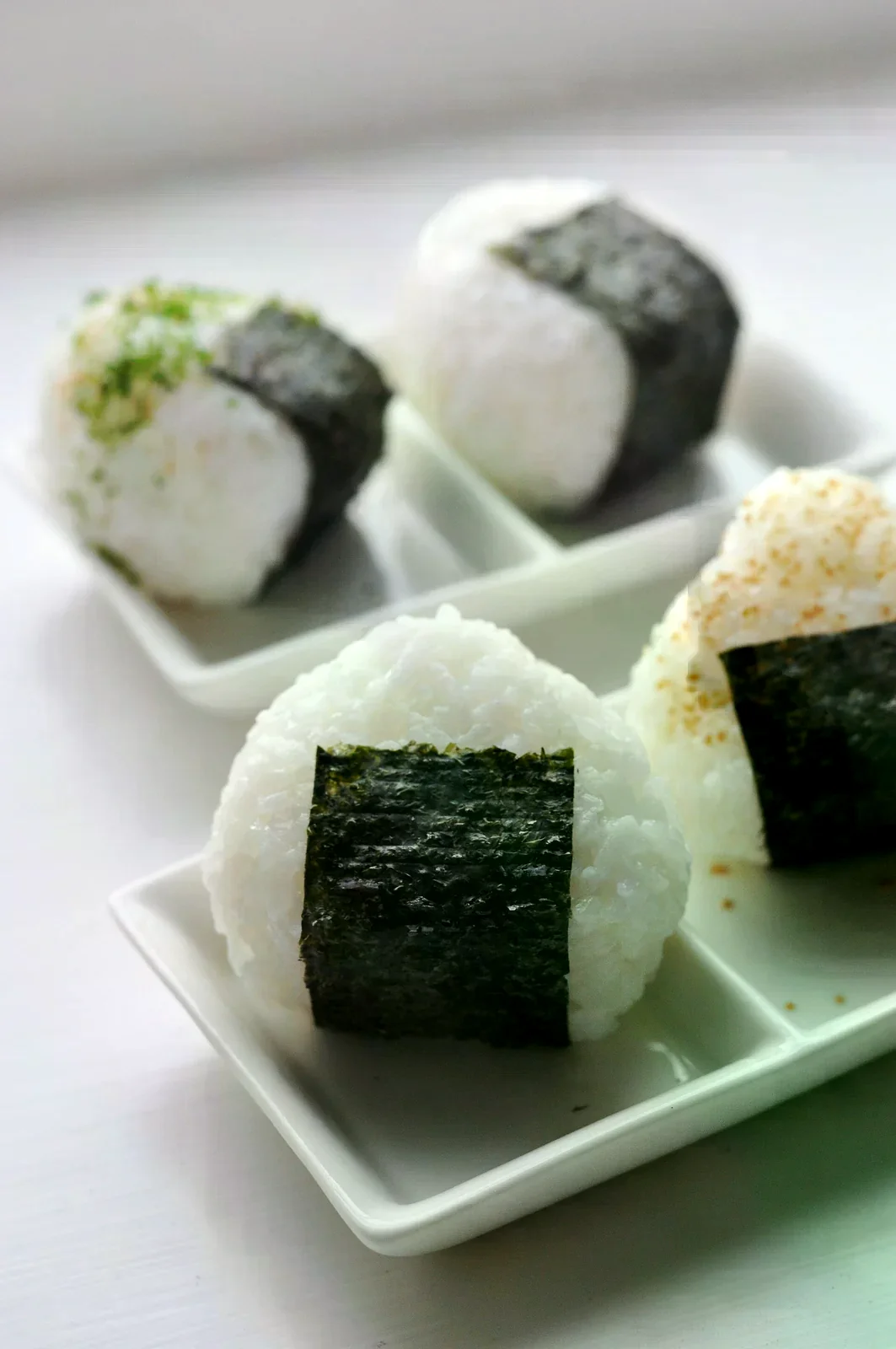 四顆白米飯糰放在白色分格餐盤上，外側裹著海苔，其中兩顆表面撒了香鬆