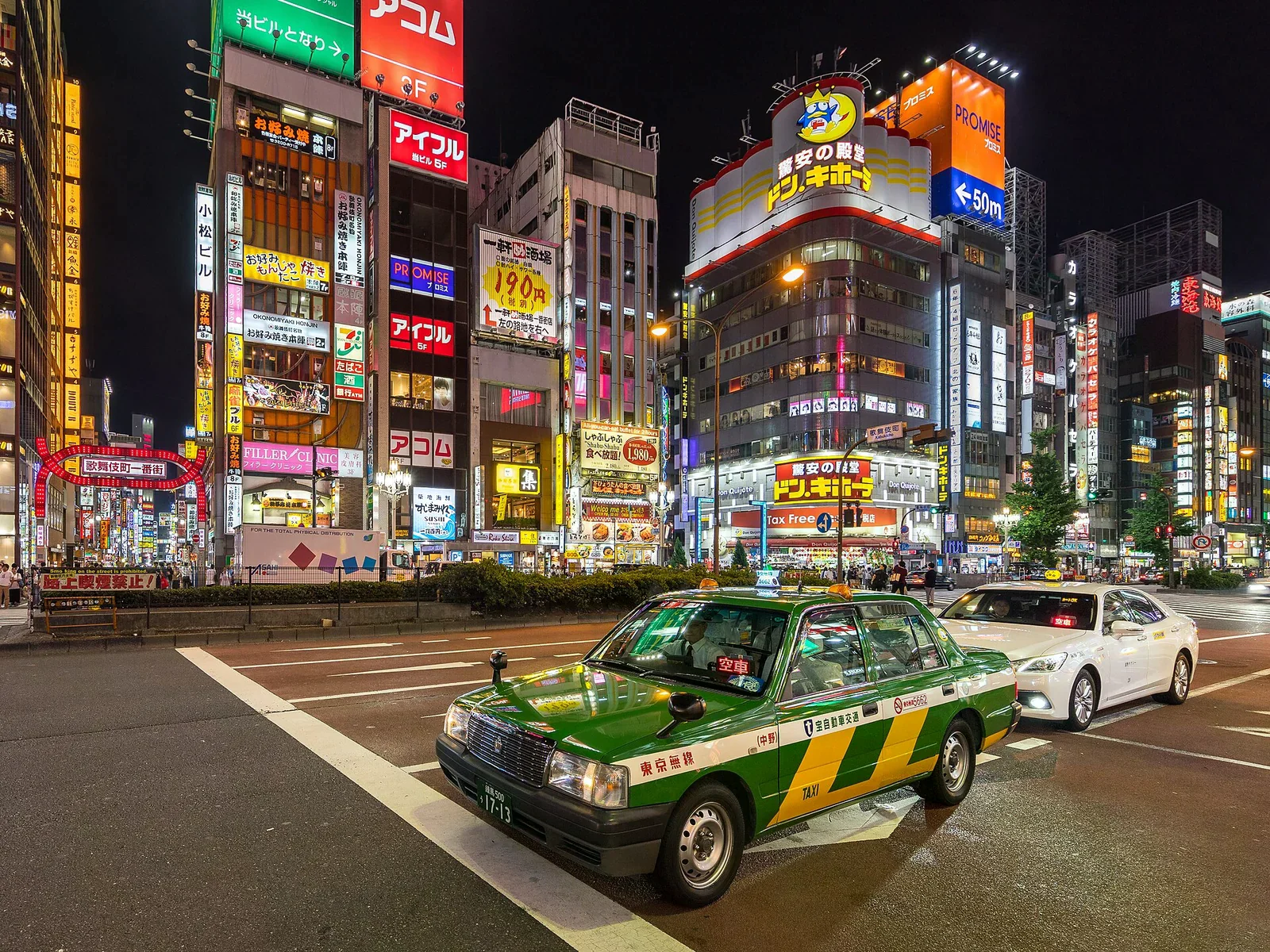 新宿靖國通夜間，一輛綠黃色東京無線 Crown Comfort 空車停在霓虹招牌背景前，街道燈光繽紛。