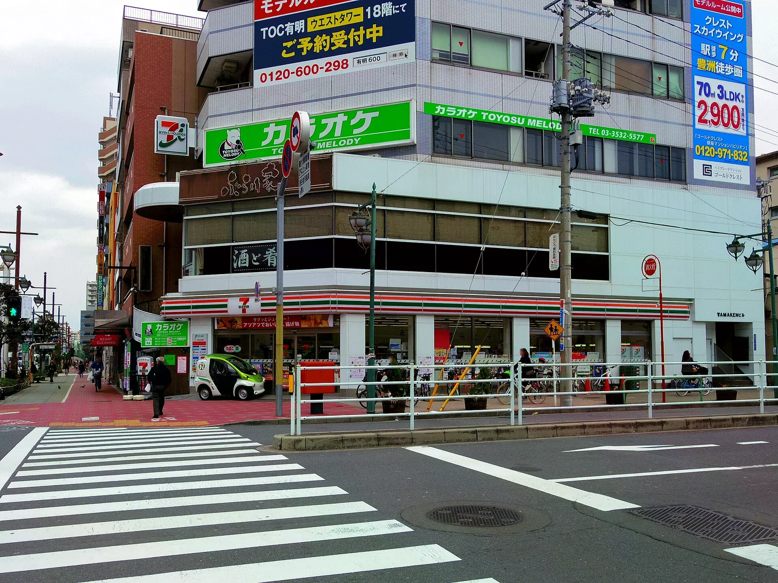 日本街角一間 7-Eleven 店面，橘綠紅條色招牌橫幅，門口有小型白色電動車與過街行人