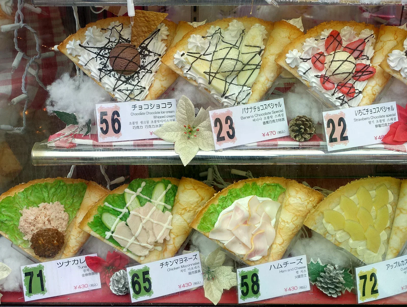 可麗餅店的展示櫃，上層是巧克力、香蕉、草莓甜點可麗餅模型，下層是生菜火腿鹹口味，前方放著中日韓英對照的價格牌