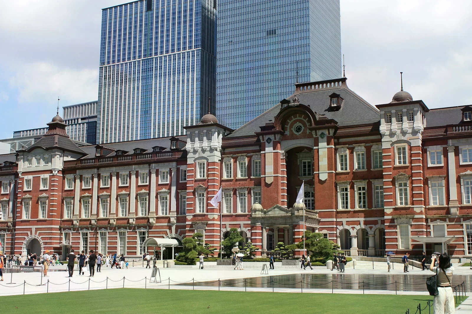 東京站丸之內側的紅磚歷史車站建築，前方是鋪了草坪與噴水池的站前廣場，後方有現代玻璃高樓