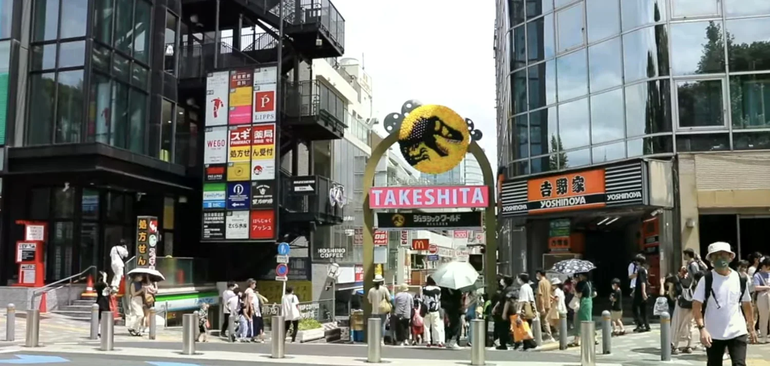 東京原宿竹下通入口拱門，粉紅色 TAKESHITA 字樣與恐龍招牌，兩旁是藥妝店與吉野家，行人撐傘通過斑馬線