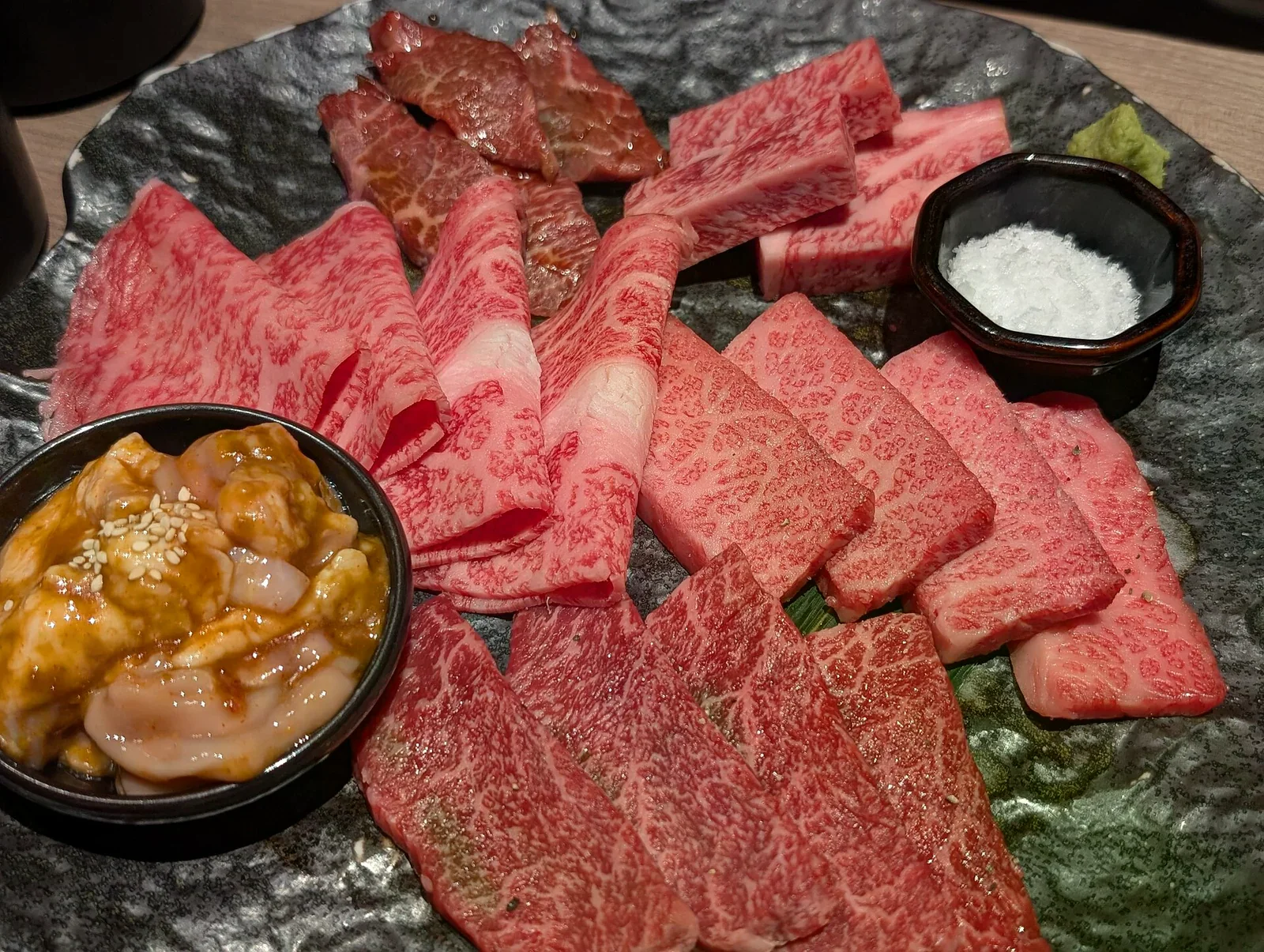 黑石盤上擺著綜合生牛肉拼盤，旁邊有鹽碗與醬料小碗