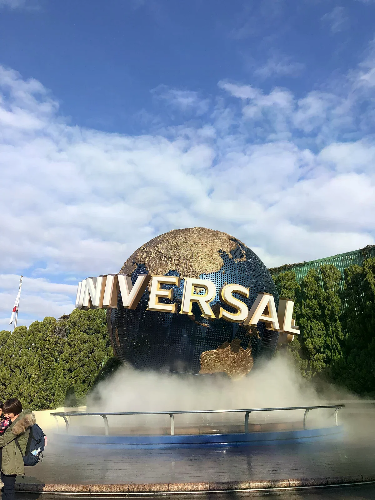 日本環球影城正門前的 UNIVERSAL 地球裝置，藍白金邊大字，底座噴著白霧，背景是藍天白雲與綠樹籬